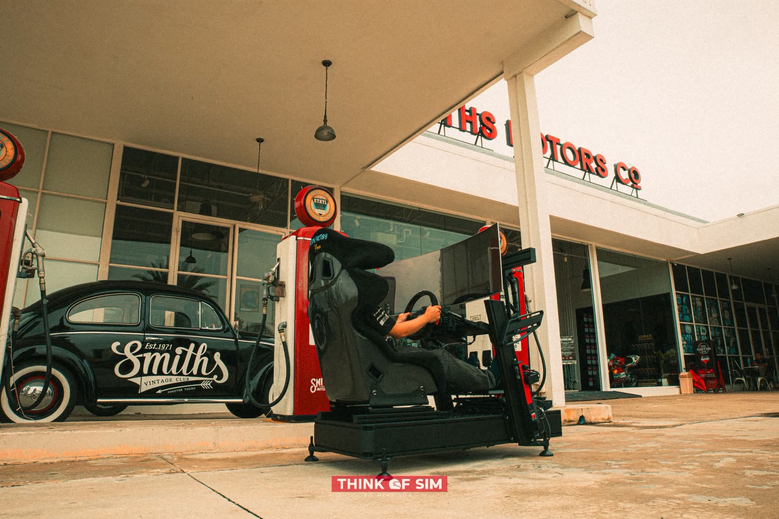 Think of Sim racing rig photographed at a retro petrol station beside a classic Beetle
